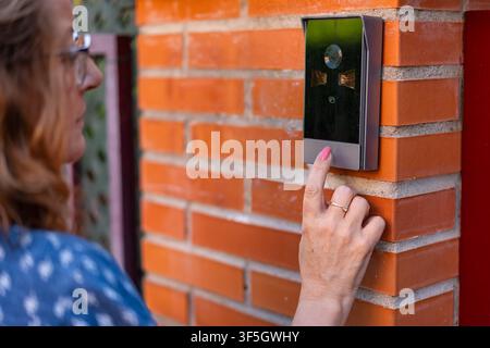 Woman pressing a modern video doorbell mounted on an orange brick entrance wall outdoor Stock Photo