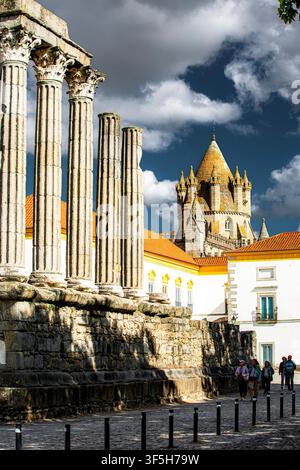 Ancient Roman columns stand tall against dramatic clouds. Stone ruins show weathered texture and historical depth. Towering church spire rises behind Stock Photo