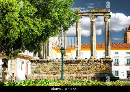 Ancient Roman columns stand tall in Evora, Portugal. Sunlit stone pillars rise beside lush green trees. Bright blue sky with fluffy clouds enhances th Stock Photo