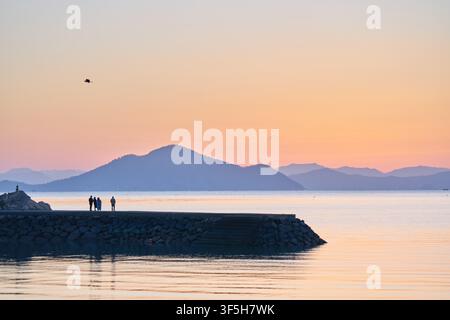 Sunrise over the sea near Sodeungseom, South Korea, with people silhouetted on a breakwater and distant mountains glowing at dawn. Stock Photo