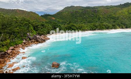 A tranquil beach scene with turquoise waves rolling onto the white sand, backed by lush, green hills and dense palm trees. Anse Lazio, Seychelles. Stock Photo
