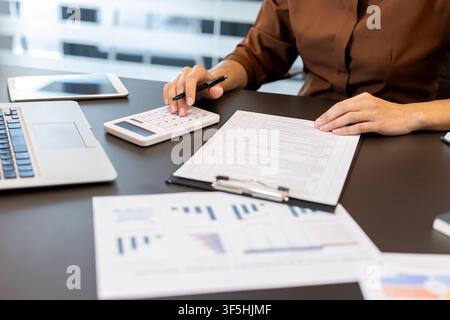Businesswoman calculating financial data using a white calculator, managing paperwork and reviewing documents with charts while working on a silver laptop on a modern office desk Stock Photo
