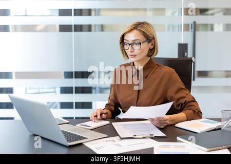Businesswoman managing finances, analyzing documents, and using a calculator with a laptop on a contemporary office desk, focusing on data and financial planning Stock Photo