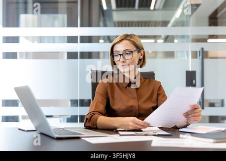 Professional businesswoman wearing eyeglasses reviewing important paperwork and concentrating on her work at a desk with a laptop, managing tasks in a contemporary corporate environment Stock Photo
