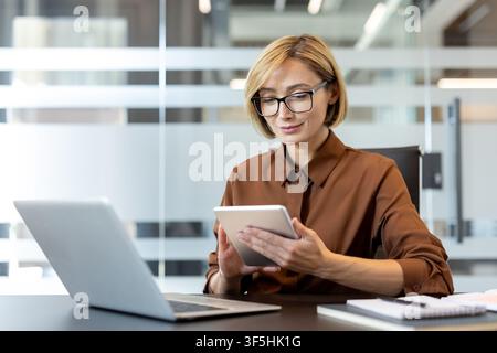 Confident businesswoman with eyeglasses using a digital tablet at her desk. Working on corporate tasks with a laptop and notebooks nearby Stock Photo