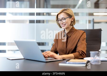 Professional businesswoman wearing eyeglasses smiling while typing on a laptop, effectively managing tasks and achieving success in a contemporary office environment Stock Photo