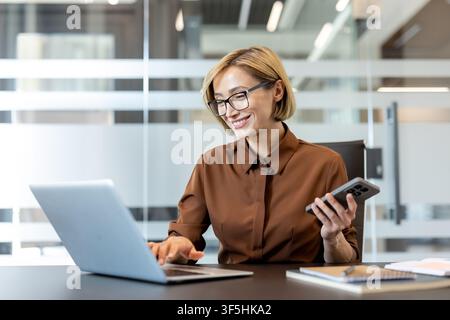 Smiling businesswoman sitting at office desk. Actively working on a laptop while holding a smartphone. Representing modern productivity. Connectivity Stock Photo