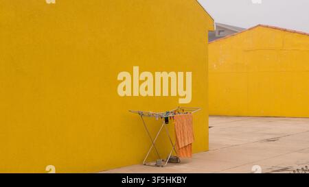 Orange Towel Drying on a Folding Rack Against a Bright Yellow Rendered Wall — Concept: Minimalist Colour Block Composition, Everyday Domestic Life Stock Photo