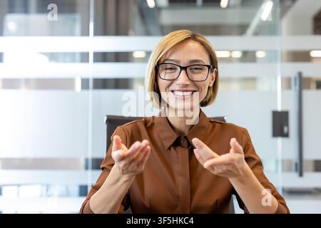 Woman wearing glasses and a brown shirt talking during a virtual meeting, communicating effectively in a modern office environment Stock Photo