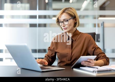 Professional businesswoman using a laptop and holding a tablet in a modern office, engaged in multitasking and managing tasks efficiently, representing productivity and corporate success Stock Photo
