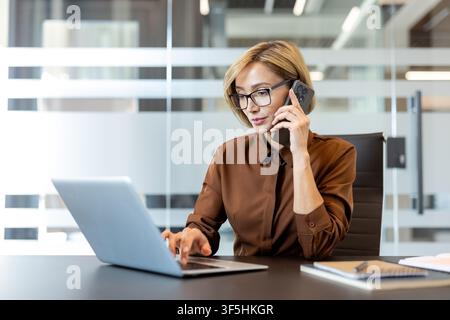 Young businesswoman managing communication and work tasks simultaneously, talking on a mobile phone while typing on a computer in a contemporary corporate environment Stock Photo
