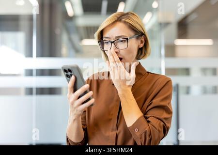 Woman standing in a modern office looking surprised while covering her mouth, receiving shocking news or an unexpected message on her mobile phone, expressing disbelief Stock Photo