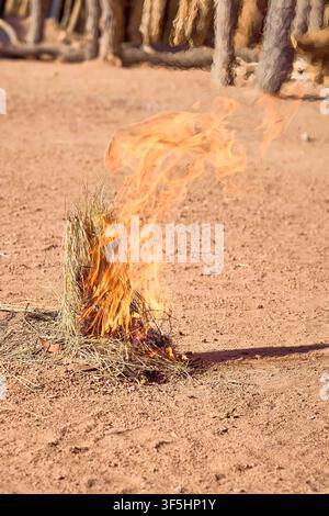 Bright orange flames erupt from a conical heap of dry grass on the sandy ground, creating a small campfire in a traditional village setup. Stock Photo