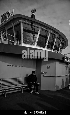 A man sits on a wooden bench aboard a passenger ferry under a stark monochrome sky Stock Photo