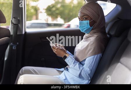 Black muslim businesswoman wearing face mask while using taxi during coronavirus pandemic Stock Photo