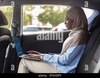 Black Muslim Businesswoman Using Laptop In Car While Going To Work Stock Photo