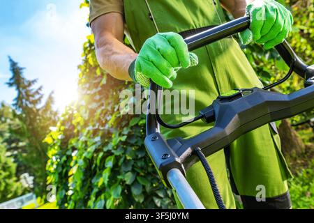 A gardener stands with a lawn mower in a sunny backyard. The man wears green gloves and overalls while trimming the grass. Lush trees and bushes surro Stock Photo