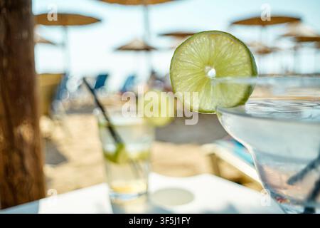 Close-up of a refreshing margarita cocktail garnished with a fresh lime slice on beach with parasols in the background. Stock Photo