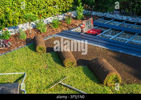 Workers are placing rolls of sod on a garden area in the afternoon. Tools are arranged nearby and landscaping features like plants and mulch are visib Stock Photo