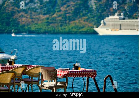 An empty dining table with a checkered tablecloth and chairs set right by the blue sea, with a large white cruise ship sailing in the background. Stock Photo