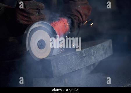Close-up of a worker’s gloved hands using an angle grinder to cut a steel beam, with bright sparks, smoke, and a rotating abrasive disc in a dark work Stock Photo