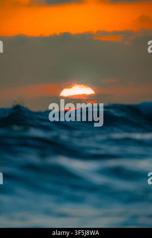 Dramatic sunset over rolling ocean waves near Santa Elena, Ecuador, with the glowing sun partly hidden behind dark clouds and a vivid orange sky above Stock Photo