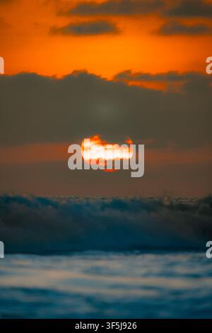 Glowing sunset descends behind dark clouds over the Pacific Ocean at Santa Elena, Ecuador, while rolling waves and foamy surf create a dramatic beach Stock Photo