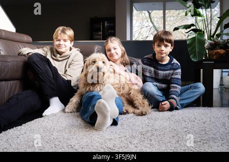Three children relax on a cozy living room carpet with their fluffy pet dog, enjoying quiet family time at home in natural light, with a sofa and plan Stock Photo