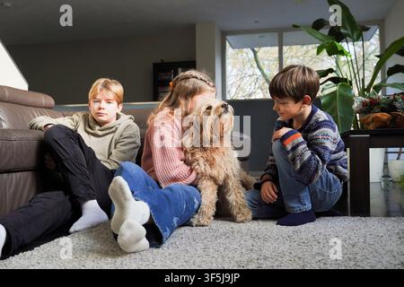 Three children relax on a carpeted living room floor with a fluffy small dog, enjoying a calm moment at home with soft daylight, sofa seating, and ind Stock Photo