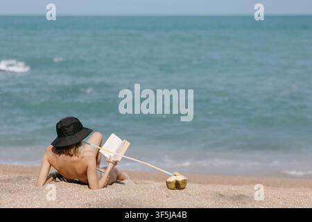 Young woman relaxes on a sandy tropical beach in Thailand, reading a book beside the sea. Mindful slow living, wellness travel, remote lifestyle, calm Stock Photo