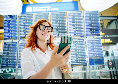 A red-haired woman in glasses is using her smartphone while standing in an airport terminal in front of a large flight information board, ready for he Stock Photo