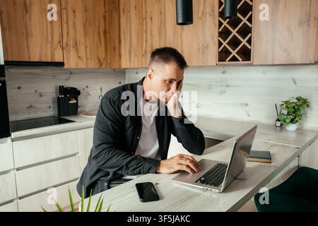 A man works remotely on his laptop in a modern kitchen setting. He appears contemplative, supporting his face with one hand while typing at a white co Stock Photo