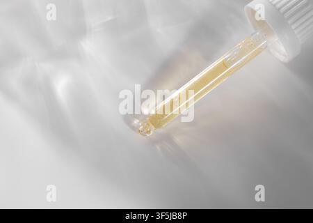 Close-up of a cosmetic pipette filled with golden gel, casting a luminous caustic effect on a soft white surface for skincare, serum, and beauty produ Stock Photo