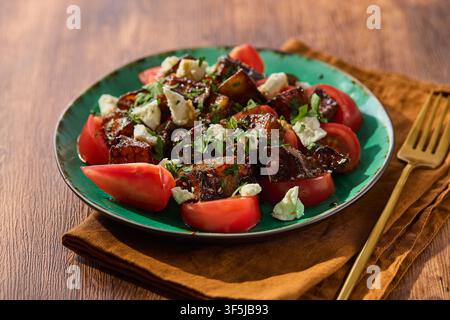 Delicious fried eggplant salad with fresh tomatoes, feta cheese, and herbs served on a green plate with a gold fork on a wooden table. Healthy vegetar Stock Photo