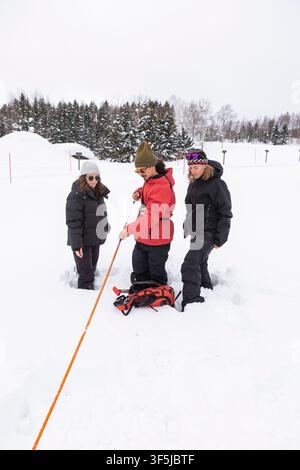 Three adults wearing winter clothing examine a long orange avalanche probe together while standing in deep snow beside a forest during a safety course Stock Photo