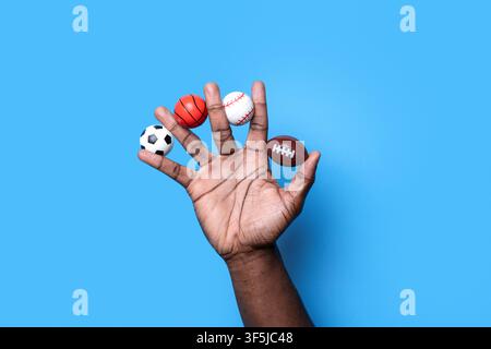 Close-up of a hand holding miniature basketball, baseball, soccer ball and American football against a bright blue background, representing sport, rec Stock Photo
