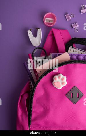 A bright pink backpack overflowing with various school and stationery supplies, including notebooks, pens, a magnifying glass, scissors, a sharpener, Stock Photo