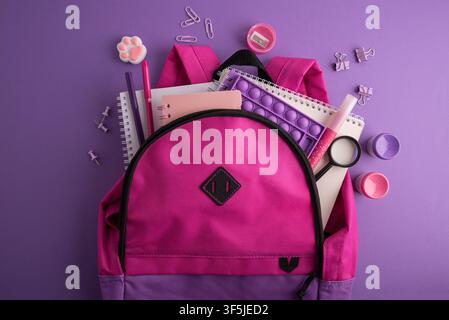 A vibrant pink backpack is overflowing with essential school supplies and stationery items, including notebooks, pens, a fidget toy, and a magnifying Stock Photo