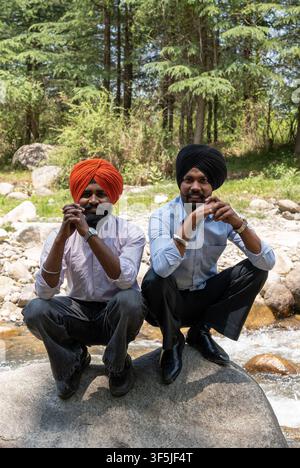 Two joyful Indian Sikh men wearing traditional turbans, both crouching down together on a river rock while smiling and looking at the camera. Stock Photo