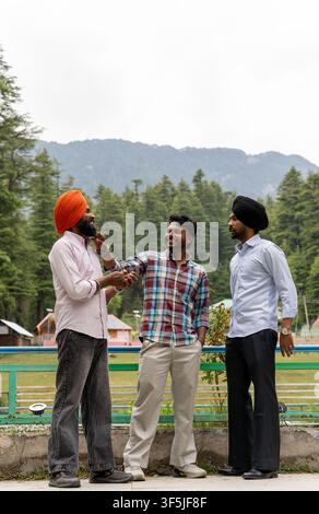 A full-length vertical shot of three Indian friends, including two Sikh men wearing traditional turbans, standing outdoors against a mountain. Stock Photo