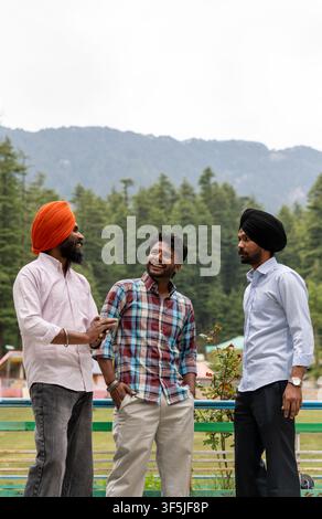 A three-quarter vertical shot of three Indian male friends, including two Sikh men wearing traditional turbans, standing outdoors against a mountain. Stock Photo