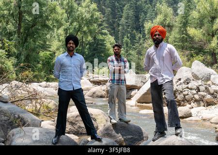 A full-length shot of three Indian friends, including two Sikh men wearing traditional turbans, standing outdoors on rocks in a river. Stock Photo