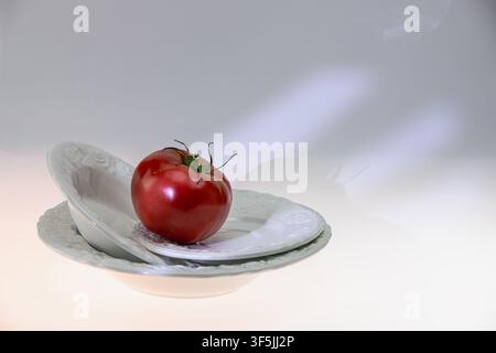 Horizontal studio still life of a red tomato with a ceramic cup on a saucer in warm light with a soft gentle shadow on a warm peach-toned background. Stock Photo