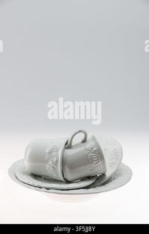 Minimal studio still life of a single white ornate ceramic cup lying on its side on a saucer against a grey background. The unexpected resting positio Stock Photo
