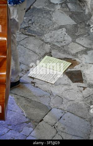 A high-angle perspective shot of an open book of sheet music lying flat on a rustic, irregular stone pavement. A portion of a musician's leg in a light-colored trouser and a black shoe, and the edge of a wooden double bass, are visible on the left side of the frame. Stock Photo