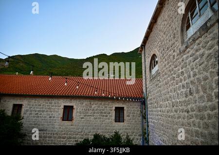 A picturesque architectural view of historic stone buildings featuring traditional orange terracotta roof tiles, small windows, arched upper windows, and string lights, set against a steep green mountain slope and blue sky. Stock Photo