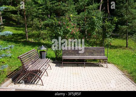 A bright, outdoor photograph showing a resting area in a park. Two empty wooden benches with ornate metal frames are positioned on a light-colored pav Stock Photo