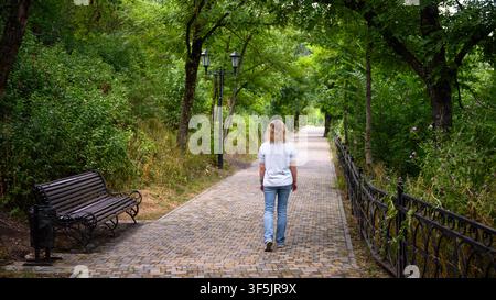 A woman with blonde curly hair is walking away from the camera down a paved path in a lush green park. She is wearing a light-colored t-shirt and blue Stock Photo