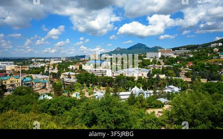 An elevated panoramic view of a city surrounded by lush green trees and rolling hills under a bright blue sky with white clouds. The cityscape feature Stock Photo