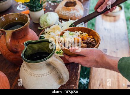 Person Serving Traditional Medieval Food From Wooden Bowl With Utensil Rustic Feast Banquet Meal Preparation Historical Reenactment Culinary Heritage Stock Photo